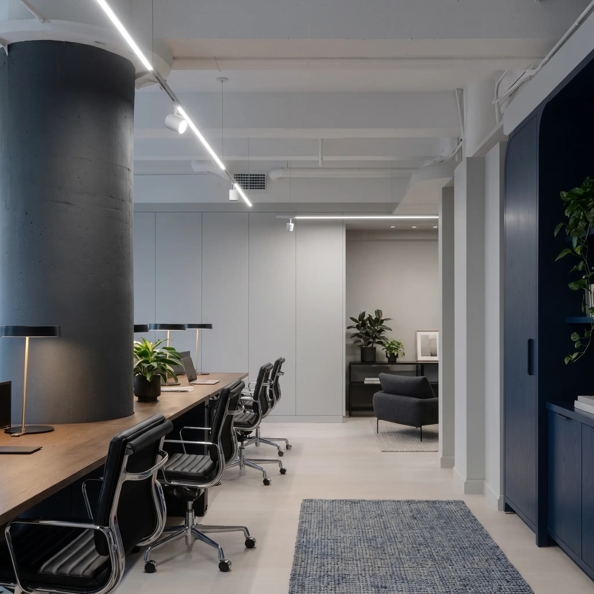 A quiet modern office desk pod with task lighting, leather chairs, indoor plants, and a lounge area beyond.